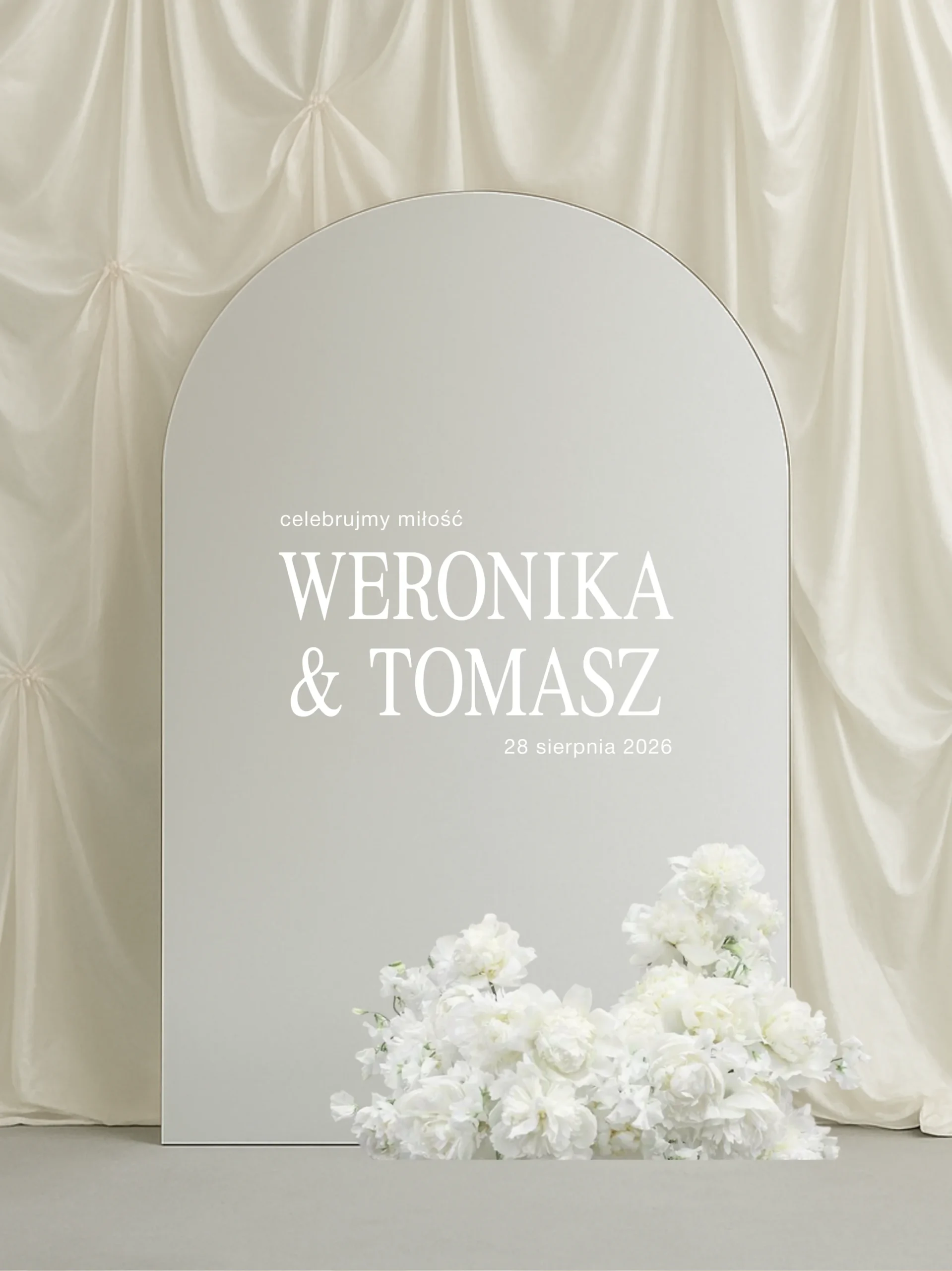 Owalne lustro ustawione pionowo na tle kremowej, udrapowanej tkaniny, z minimalistyczną białą naklejką tekstową na powierzchni lustra. U dołu lustra dekoracja z białych kwiatów; całość w jasnej, eleganckiej estetyce fine art.