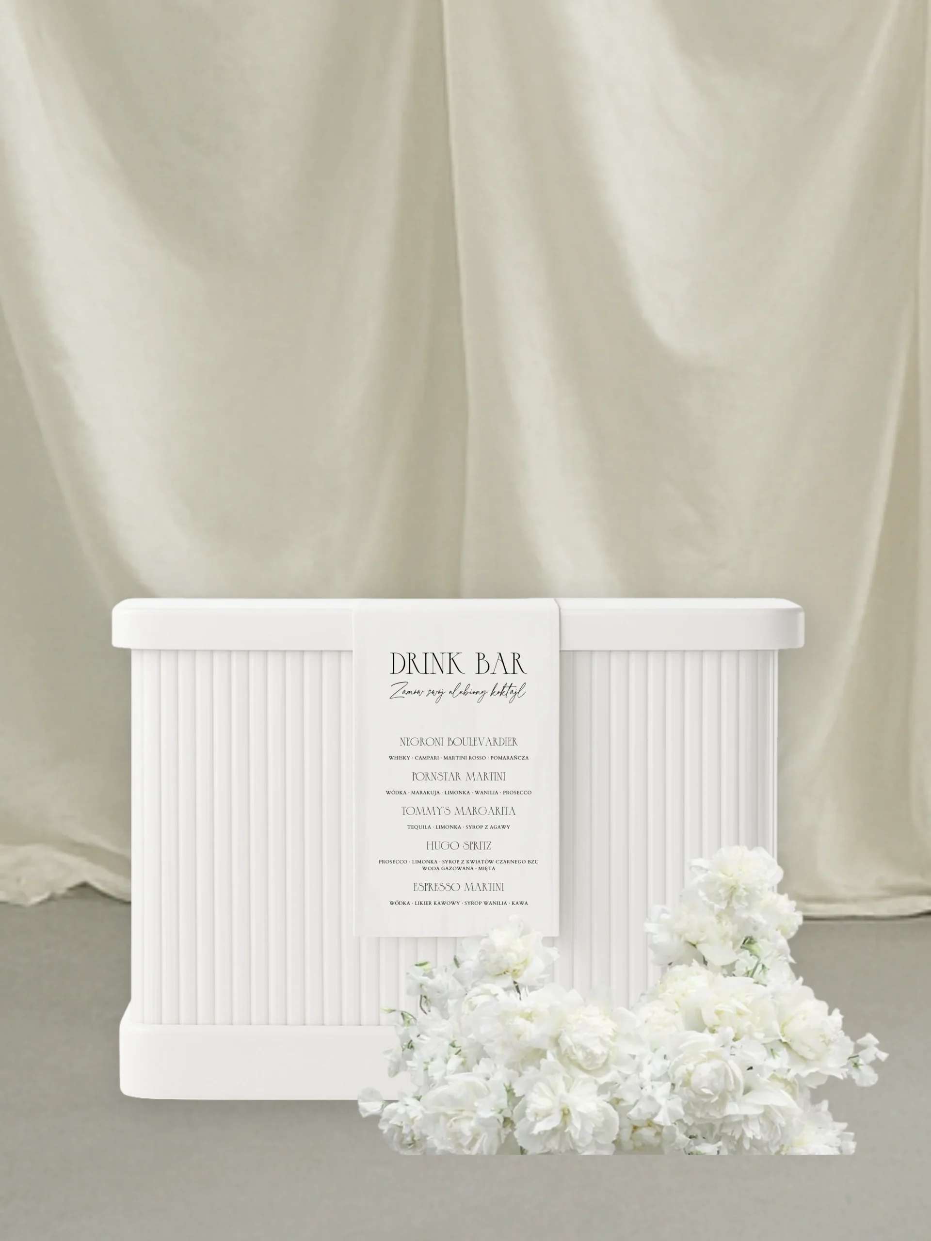 Minimalistyczna aranżacja menu drinków na jasnym tle: biała, ryflowana lada barowa ustawiona centralnie, z przodu zawieszona wąska karta „Drink Menu” z czarnym, eleganckim krojem pisma i listą koktajli. Przed ladą leży niska kompozycja z białych kwiatów o miękkiej, puszystej strukturze. Tło stanowi kremowa, miękko udrapowana tkanina, która nadaje scenie subtelny, luksusowy charakter w estetyce fine art. Całość utrzymana w jasnej, neutralnej palecie barw – bieli, écru i delikatnych beżów.